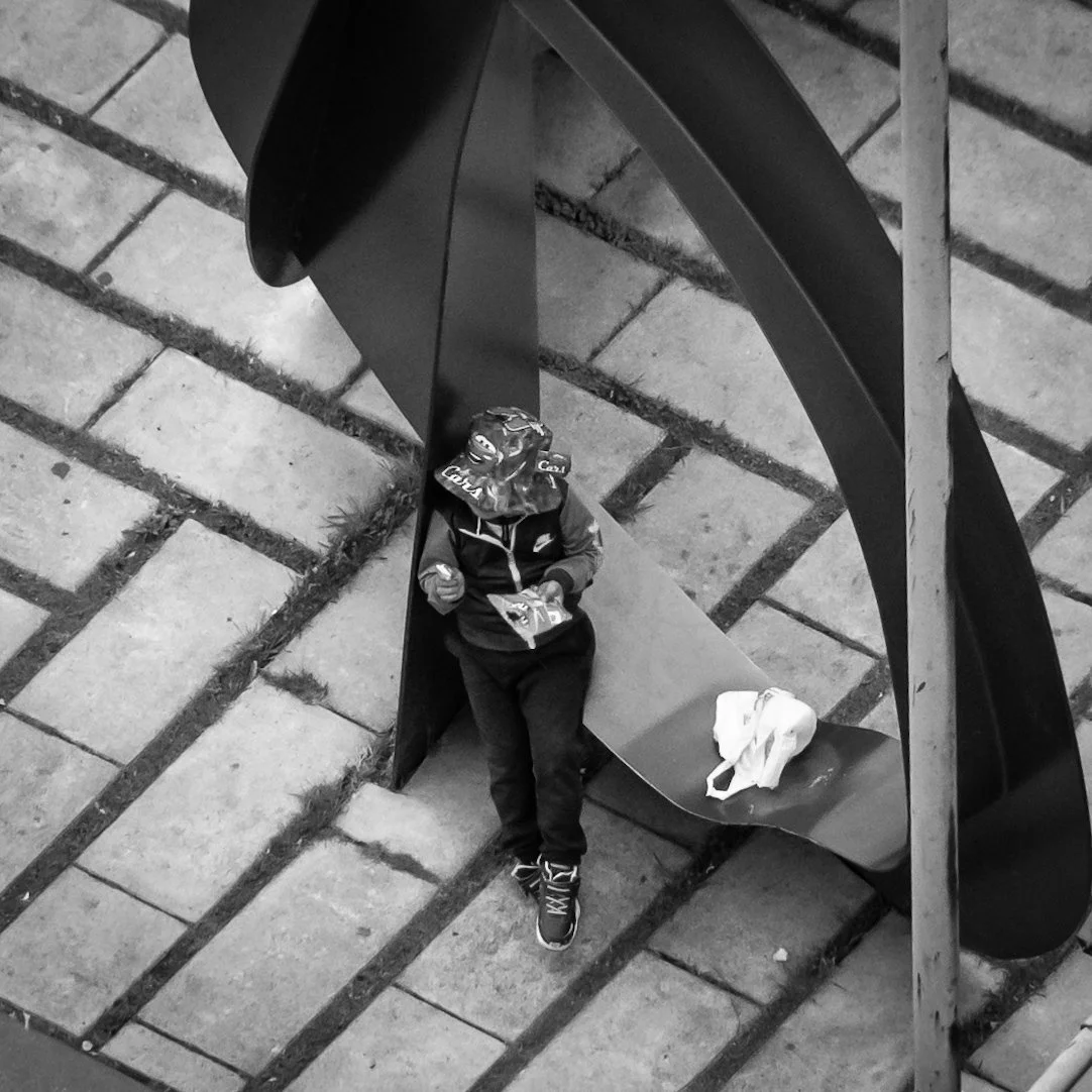 A person wearing a helmet and jacket sitting on a bench under a modern, abstract metal sculpture on a paved outdoor area, with a white bag placed on the bench next to them.