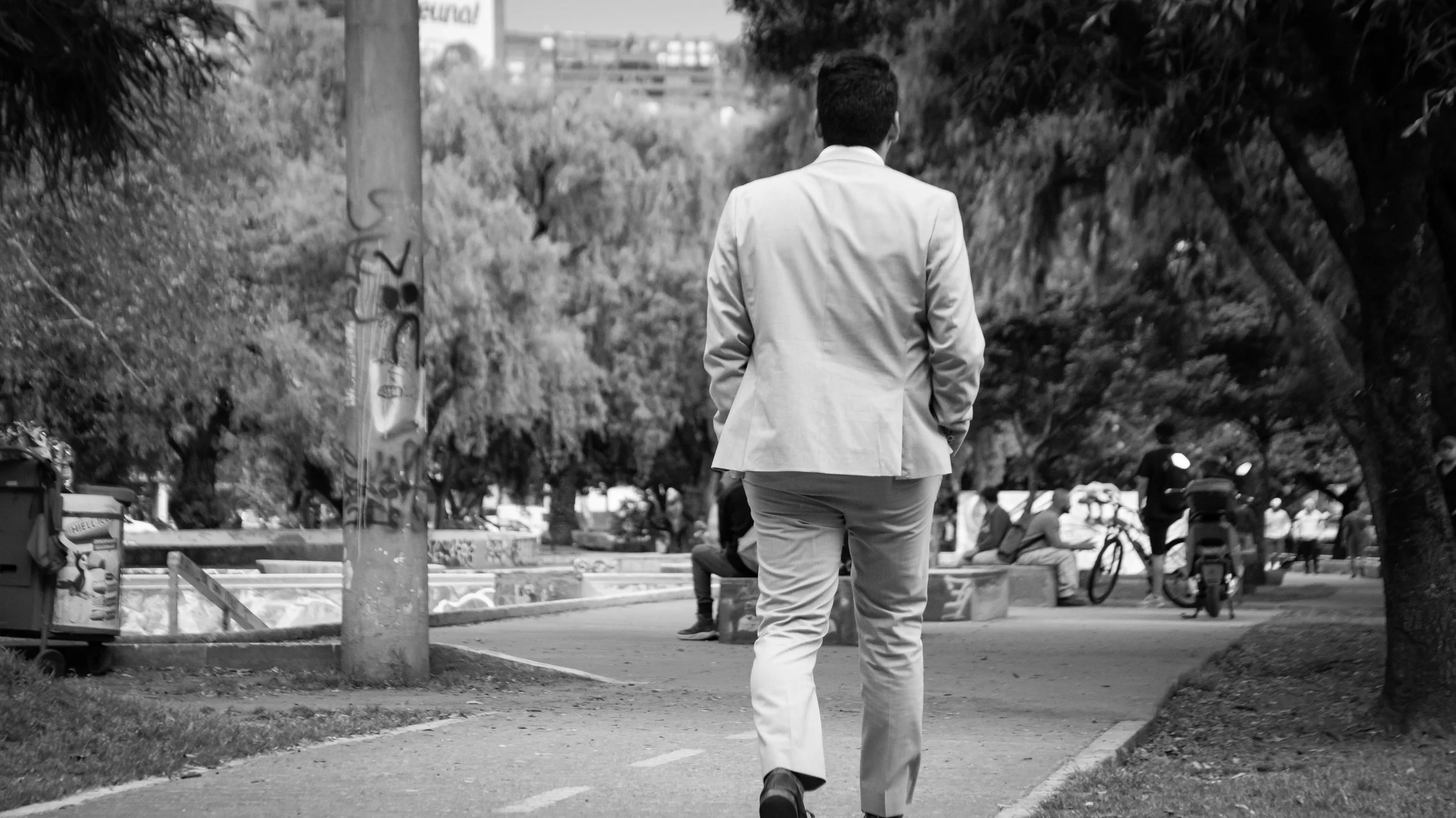 A man in a suit walking away in a park with trees, benches, and people in the background.