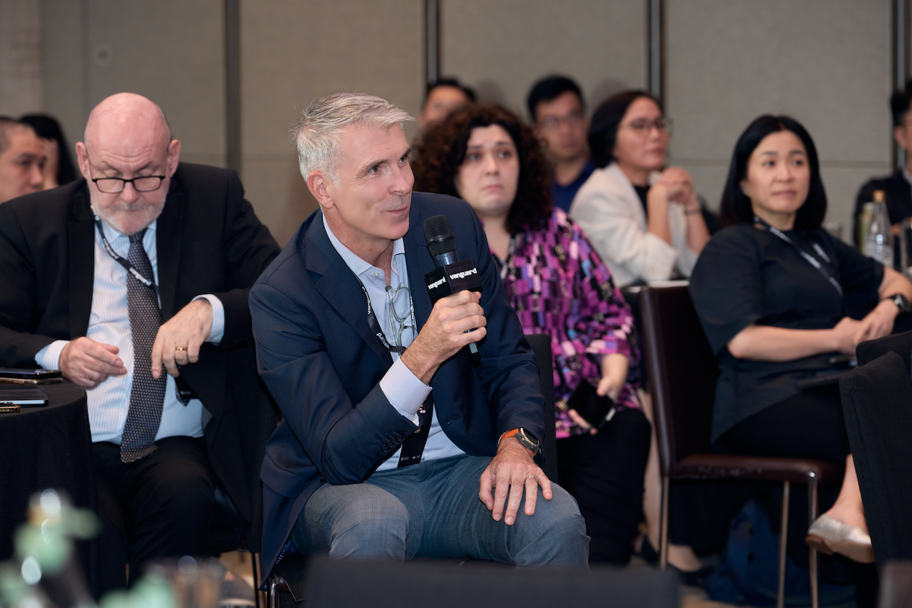 A man in a suit holding a microphone speaking at a conference with an audience.