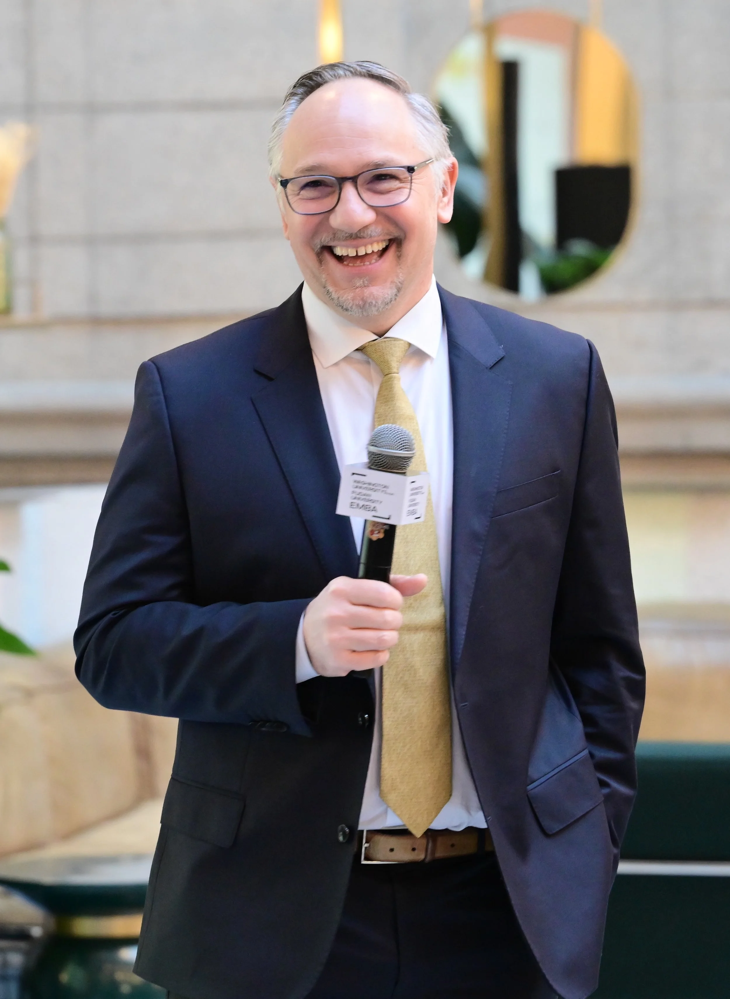 A man in a suit and tie smiling while holding a microphone at an indoor event.