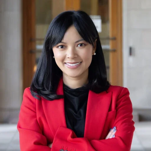 A woman with black hair wearing a red blazer and black top, smiling with her arms crossed, standing indoors with a wooden cabinet in the background.