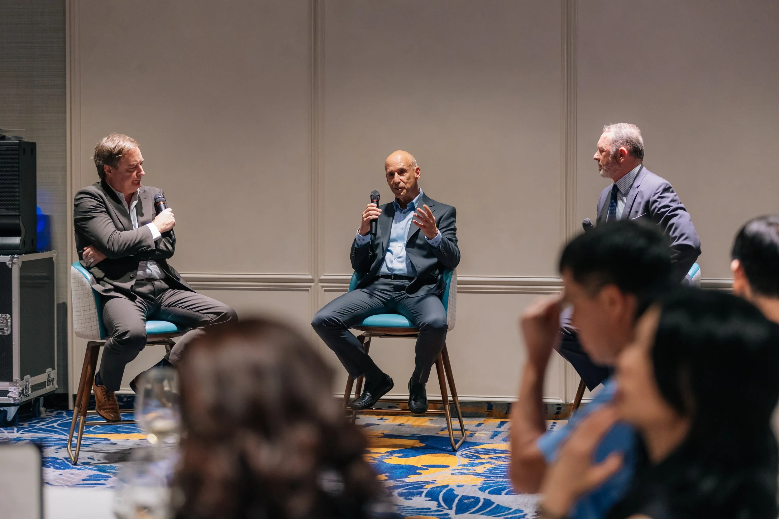 Three men are sitting and speaking in a panel discussion at a conference. They are holding microphones and engaging in conversation.