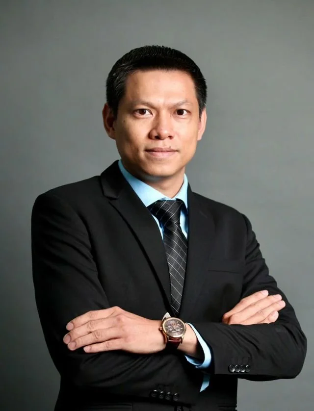 A professional man in a black suit and tie posing with arms crossed, standing against a gray background.