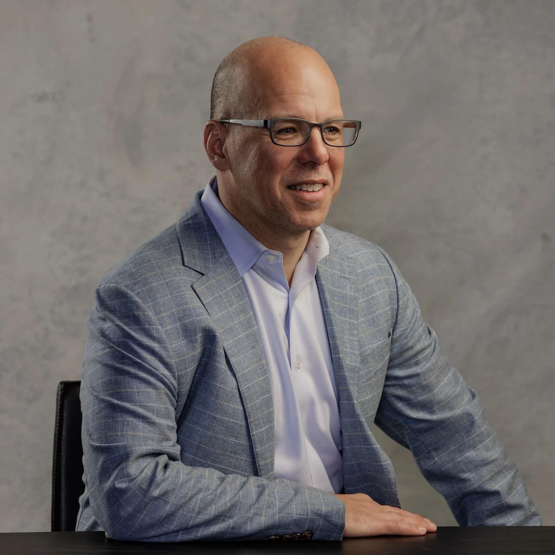 A man with glasses and a bald head sitting at a table, wearing a light blue checkered blazer and a white shirt, with a gray textured background.