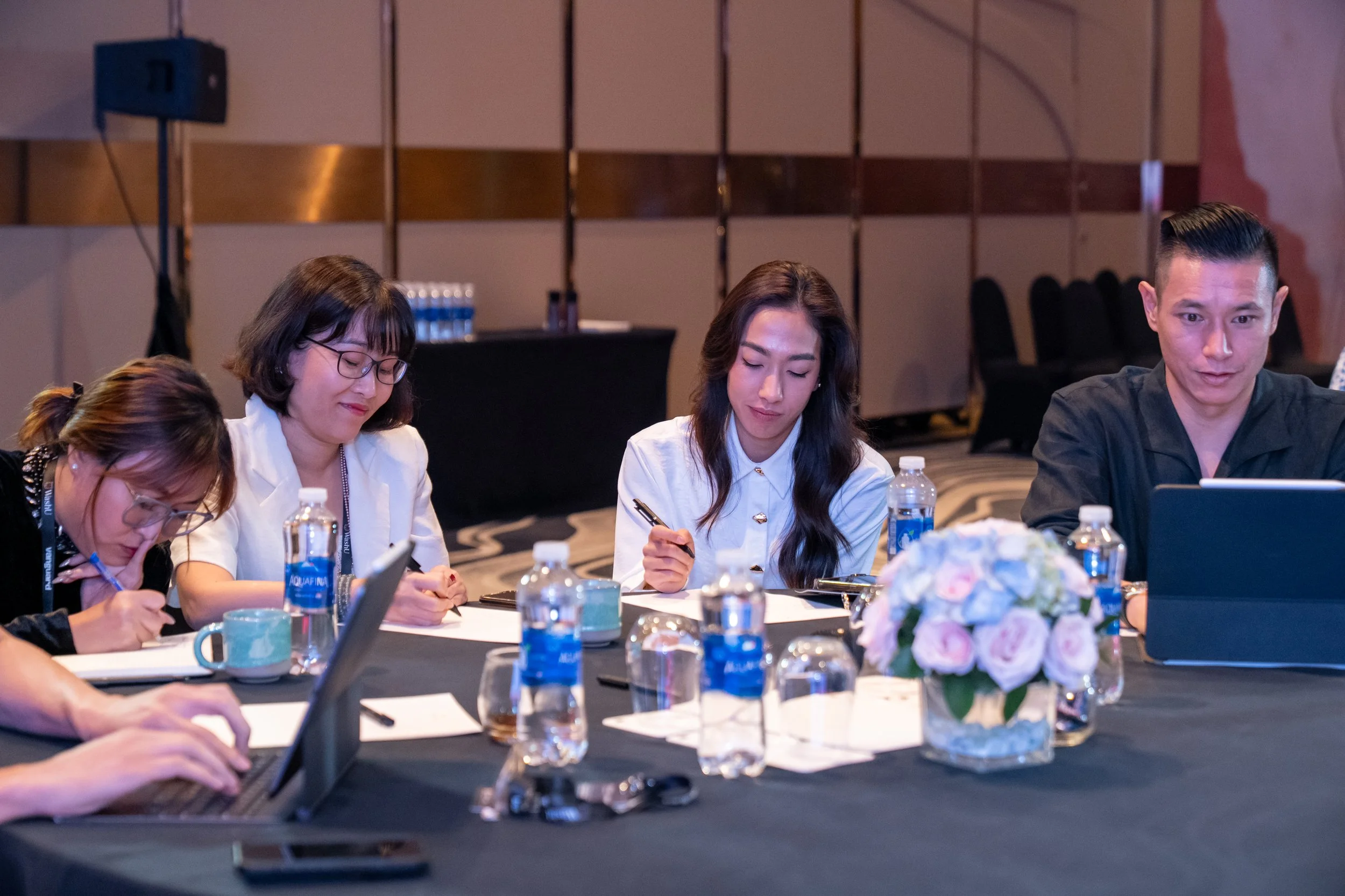 People sitting at a conference table writing notes, with bottled water, a floral centerpiece, and notebooks on the table.