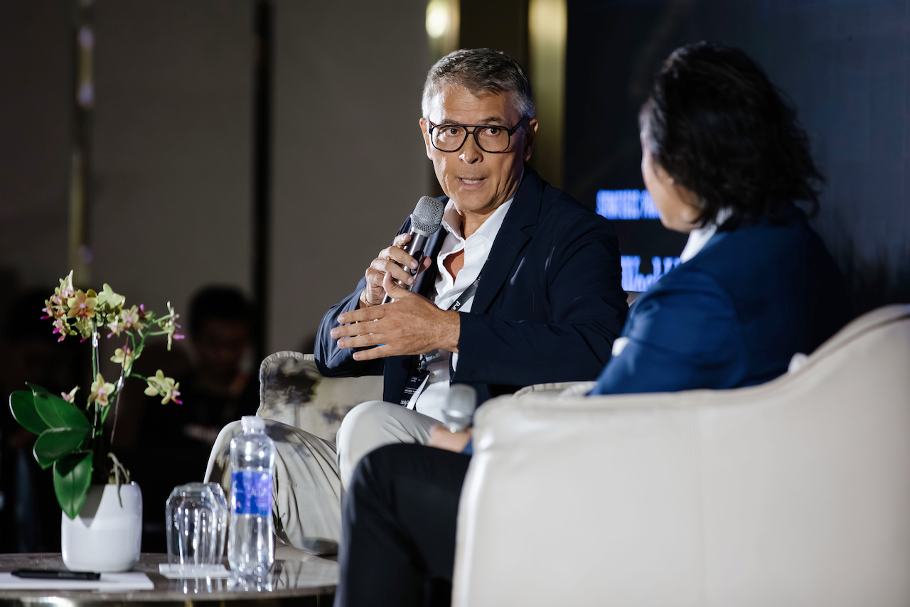 A man with glasses and gray hair, wearing a dark blazer and white shirt, speaking into a microphone during a discussion or interview at an event. He is seated on a white chair next to a woman with dark hair, wearing a blue blazer. There is a table with a potted orchid, a water bottle, and glasses in front of them.