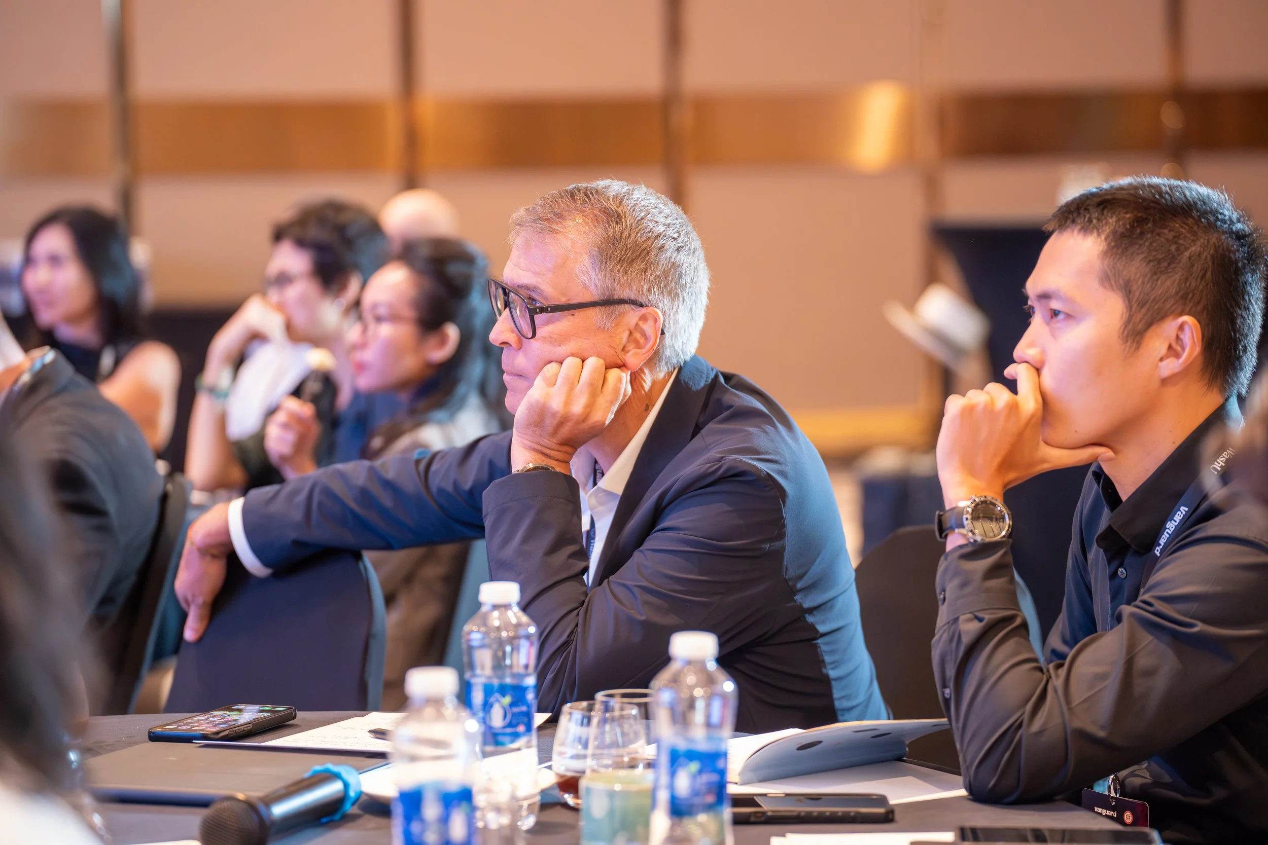 A diverse group of people attending a conference or seminar, sitting at a table, listening attentively, with water bottles and electronic devices in front of them.