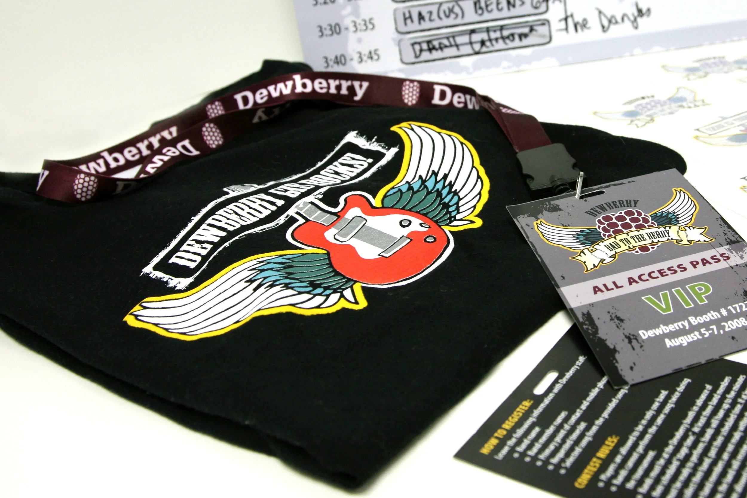 Black T-shirt with logo featuring a red berry with wings, a guitar, and the text 'Dewberry'. A VIP access pass and lanyard with the same logo are placed on the shirt, and a schedule or program is in the background.
