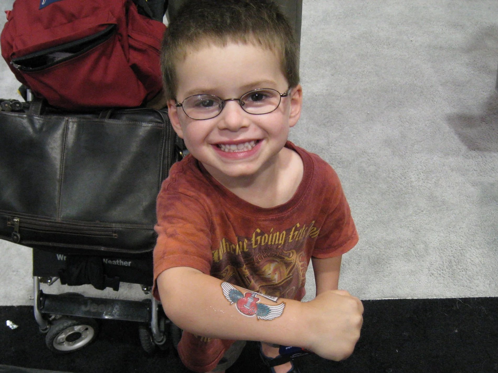Young boy with glasses smiling at the camera, showing a temporary tattoo on his arm, standing next to a suitcase. The background features carpeted flooring and parts of luggage and a backpack.