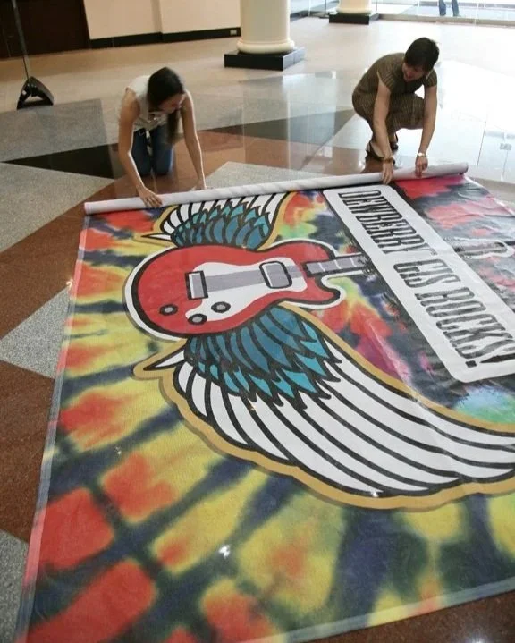 Two people laying a large, colorful banner on the floor that features a winged guitar with a red body and blue wings, and text that reads "Guitar Hero".
