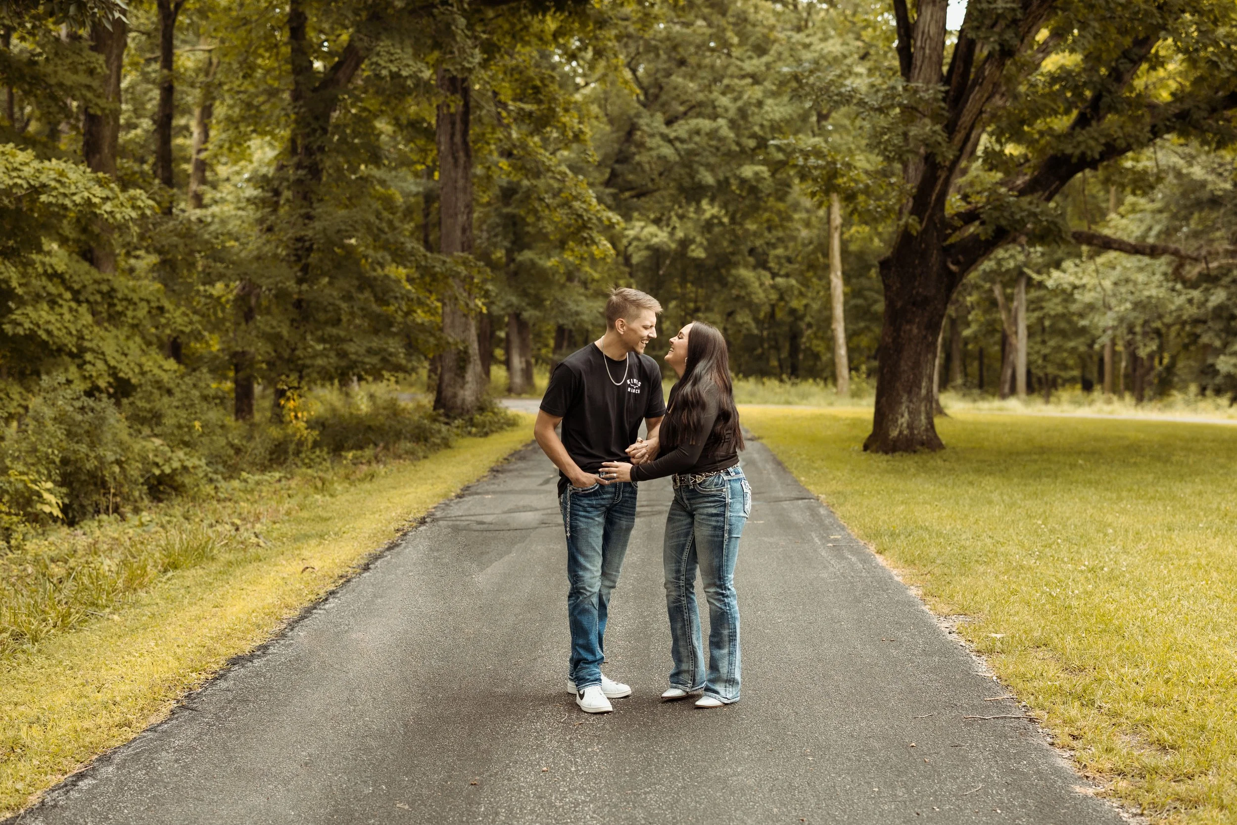 A young couple holding hands and smiling at each other while standing on a paved pathway in a lush green park with tall trees.