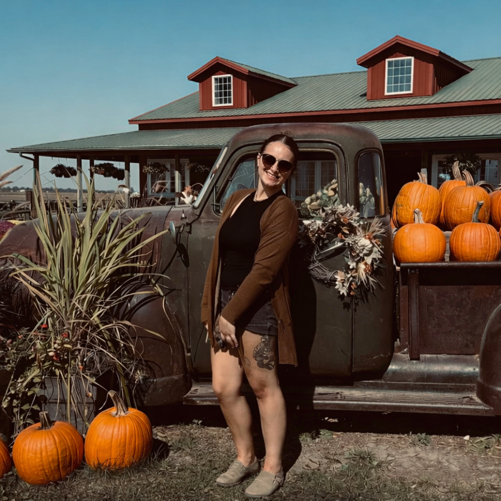 A woman with sunglasses, wearing a black shirt, a brown jacket, and denim shorts, stands next to an old black truck filled with pumpkins. The truck is parked outside a red-roofed building with a porch, under a clear blue sky.