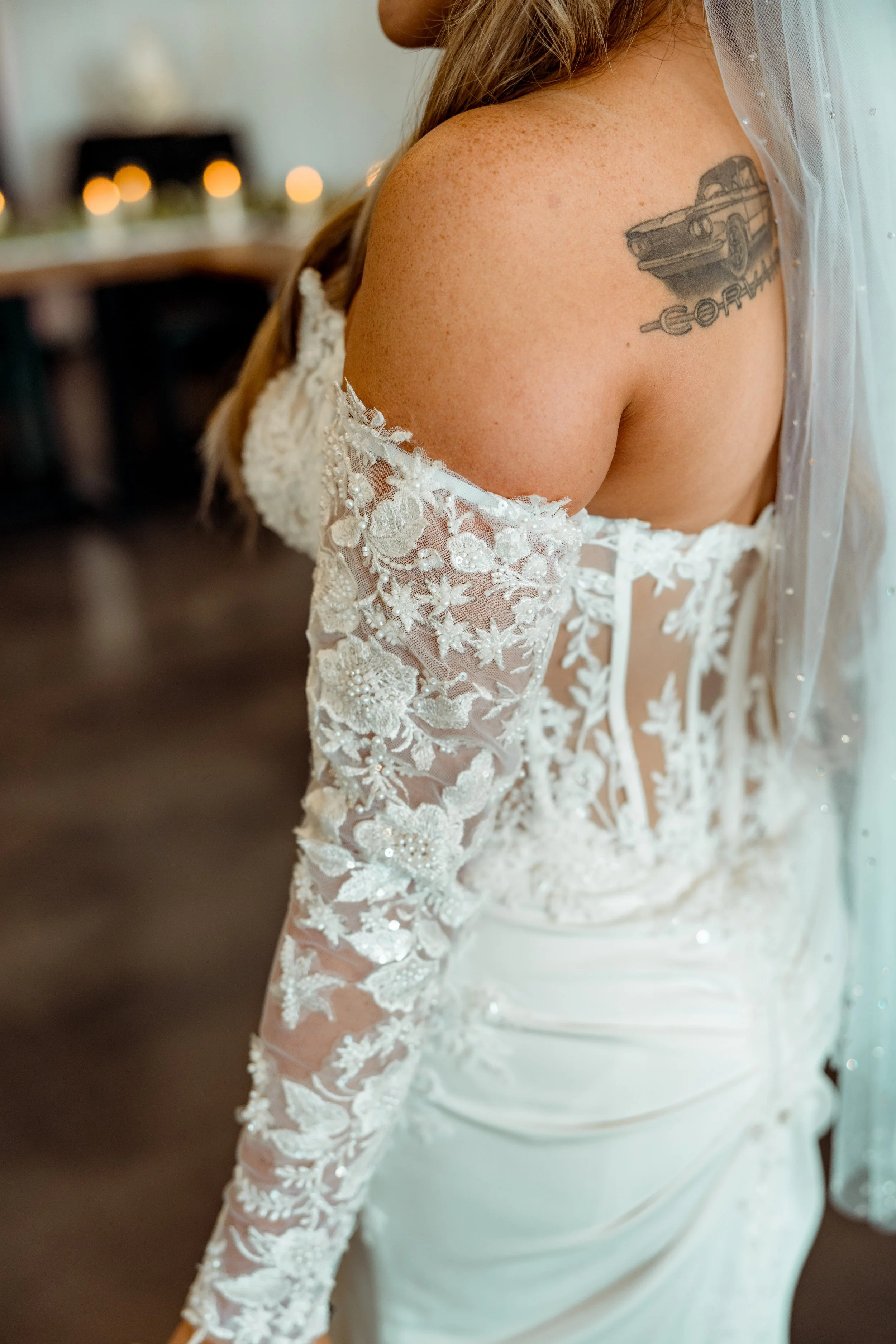 Close-up of a bride's shoulder and arm, showing a tattoo of a car,  wearing a white lace wedding dress with floral embroidery and a sheer veil.