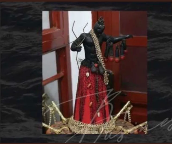 Statue noire d'une figure anthropomorphe avec un long vêtement rouge, portant un collier doré, et tenant un arc. La statue est placée sur une surface dorée ornée.