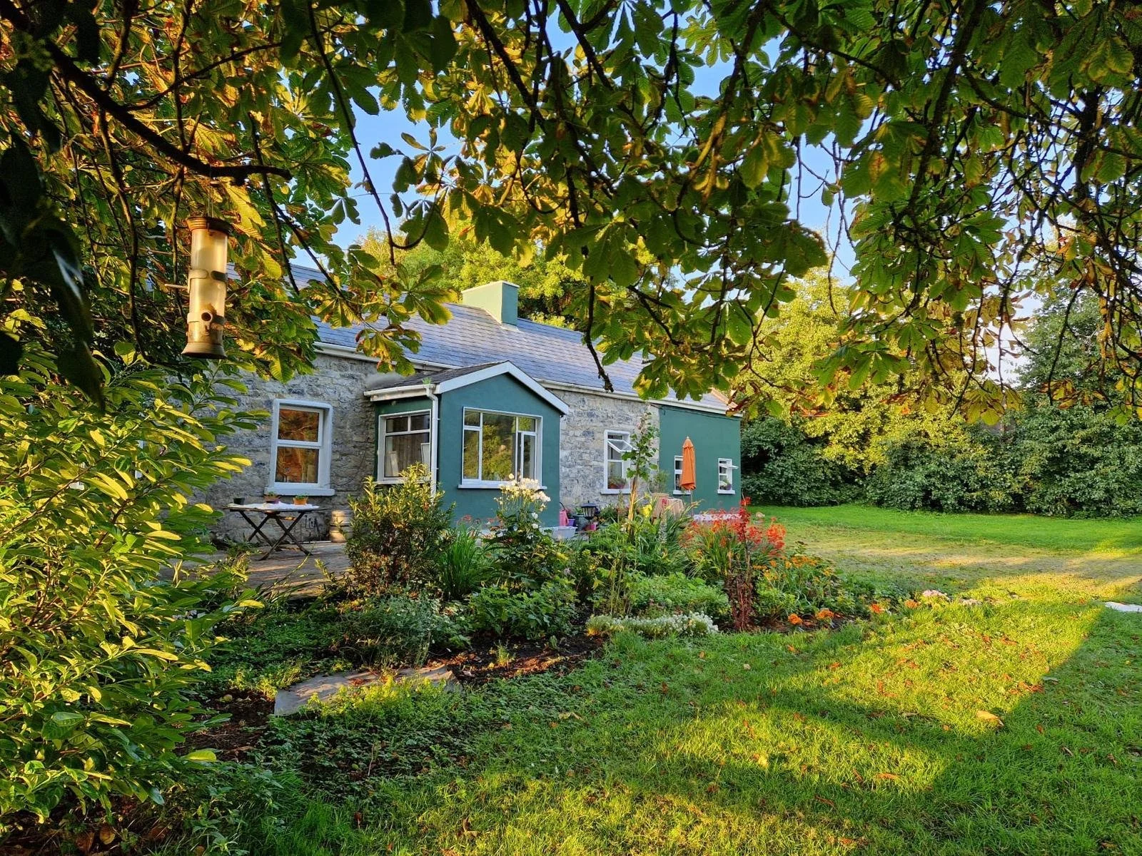 A garden with flowers and plants surrounds a small house with gray stone walls, white-framed windows, and a blue-green porch. The scene is shaded by a large tree with green leaves and a birdhouse hanging from a branch. The lawn is green and well-maintained.