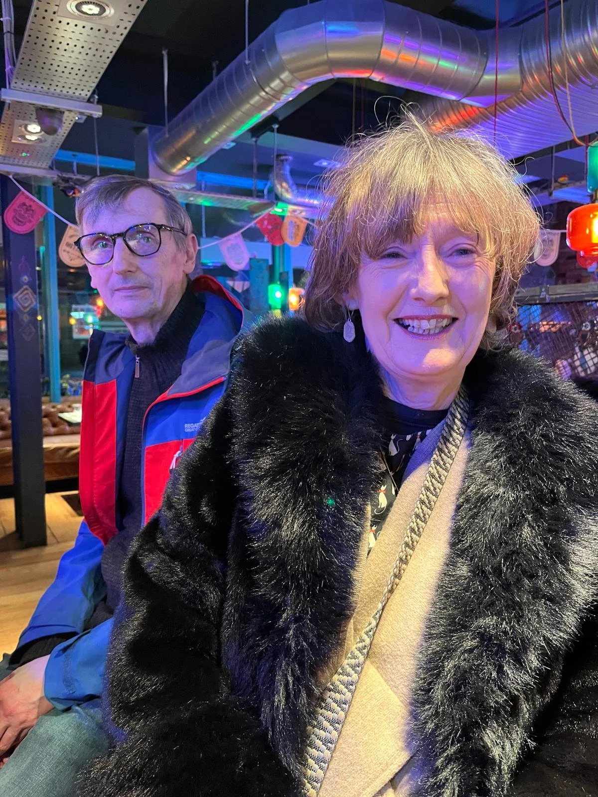Breda with short, curly blonde hair wearing a black fuzzy coat and earrings, sits next to a friend with glasses, short hair, wearing a colorful jacket, in a festive setting with colorful lights and decorations.