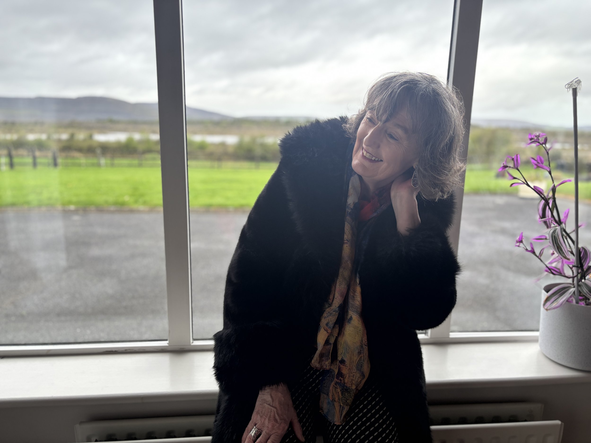 Breda smiling and standing by window with a view of an overcast sky and green fields, decorated with a potted plant with purple flowers.