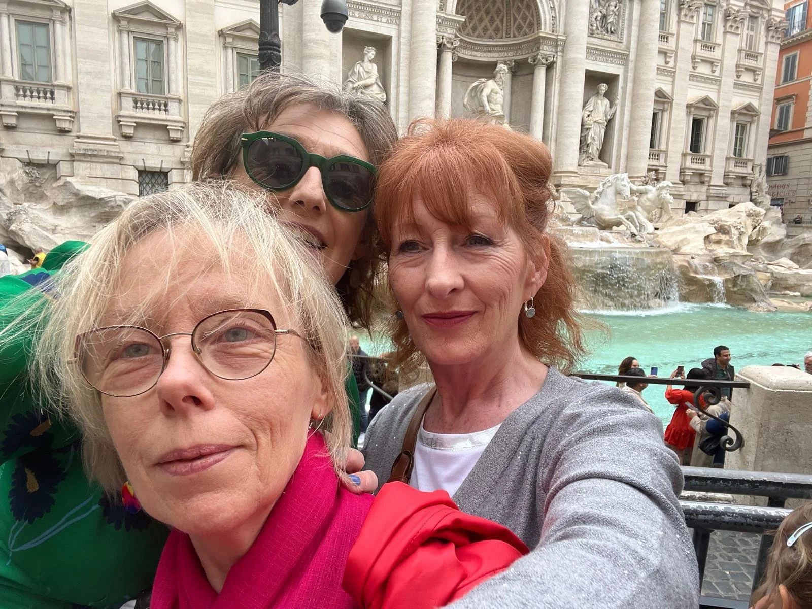 Breda and friends taking a selfie in front of the Trevi Fountain in Rome, Italy, with the fountain's sculptures and water visible in the background, and other tourists around.