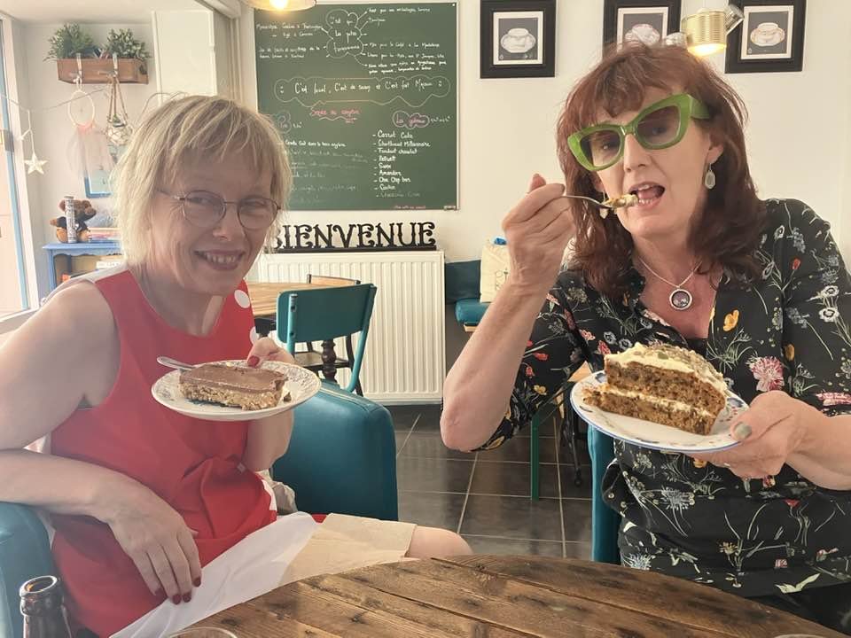 Breda and friend sitting at a table with slices of cake, one wearing glasses and a red sleeveless top, the other with green glasses and a black floral shirt, in a cafe with a chalkboard menu in the background.