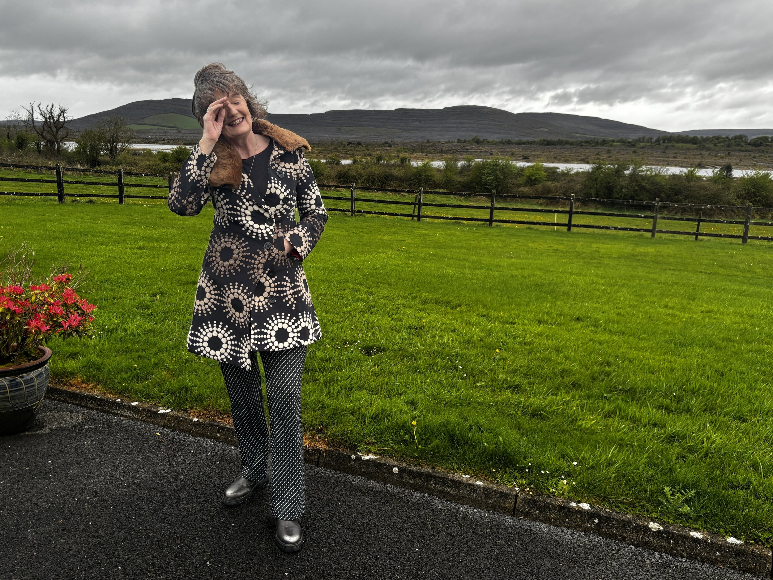 Breda standing outdoors on a paved surface, wearing a patterned coat and pants, with a scenic background of green grass, trees, a fence, and hills under a cloudy sky.