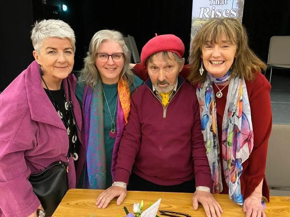 Breda and friends posing together in an indoor space, with a poster behind them that reads 'That Rises.' The man in the center is wearing a red beret and a maroon zip-up jacket, and the women are dressed in colorful clothing, smiling at the camera.