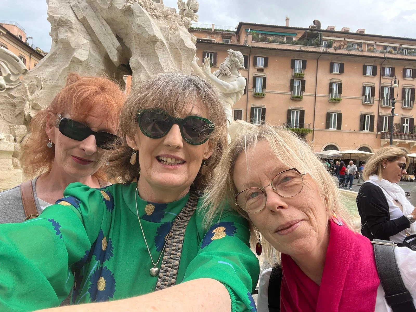 Breda and friends taking a selfie in Italy in front of a historic fountain with sculptures, with multi-story buildings in the background, and other people in the vicinity.