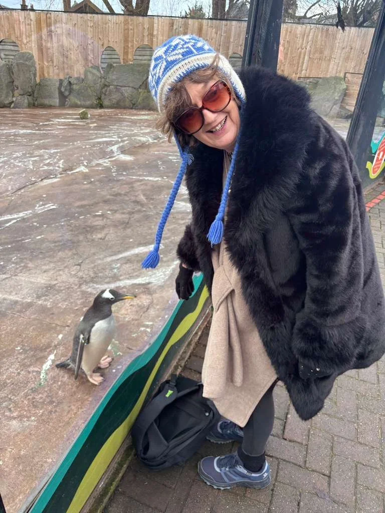Breda, wearing a blue and white knit hat, sunglasses, a black fur coat, and gray sneakers, is leaning over a glass enclosure at a zoo, smiling at a penguin standing on the ground inside the enclosure.