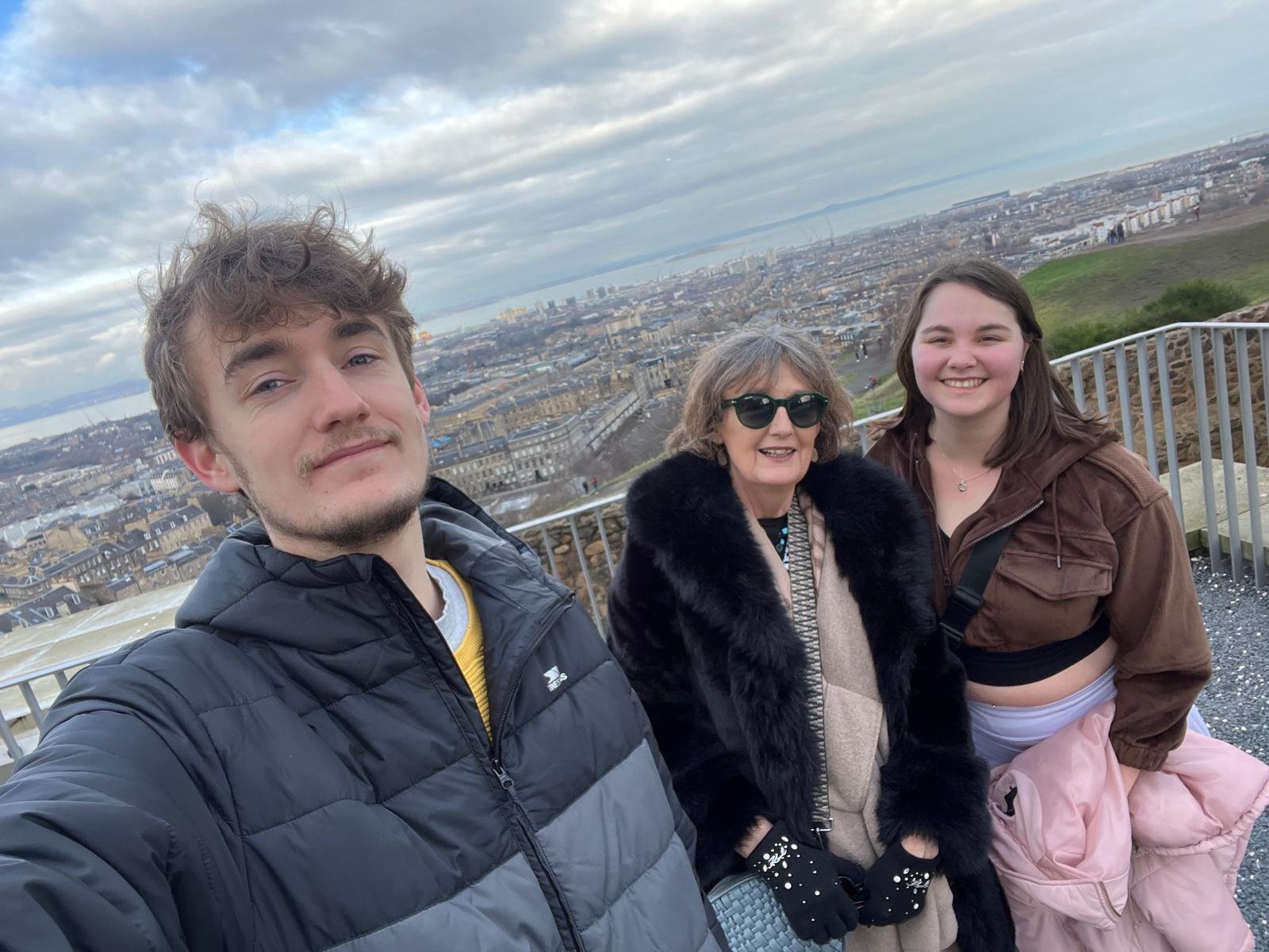 Breda, son and his girlfriend taking a selfie outdoors on a high vantage point with a cityscape and water in the background. The group includes a young man, an older woman wearing sunglasses, and a young woman with a pink jacket.