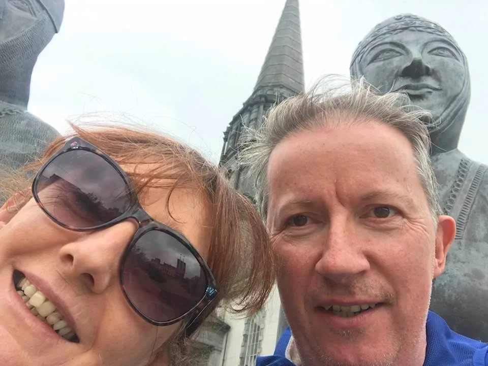 Close-up selfie of Breda wearing sunglasses and her friend, with a large sculpture of a smiling face and a church steeple in the background.