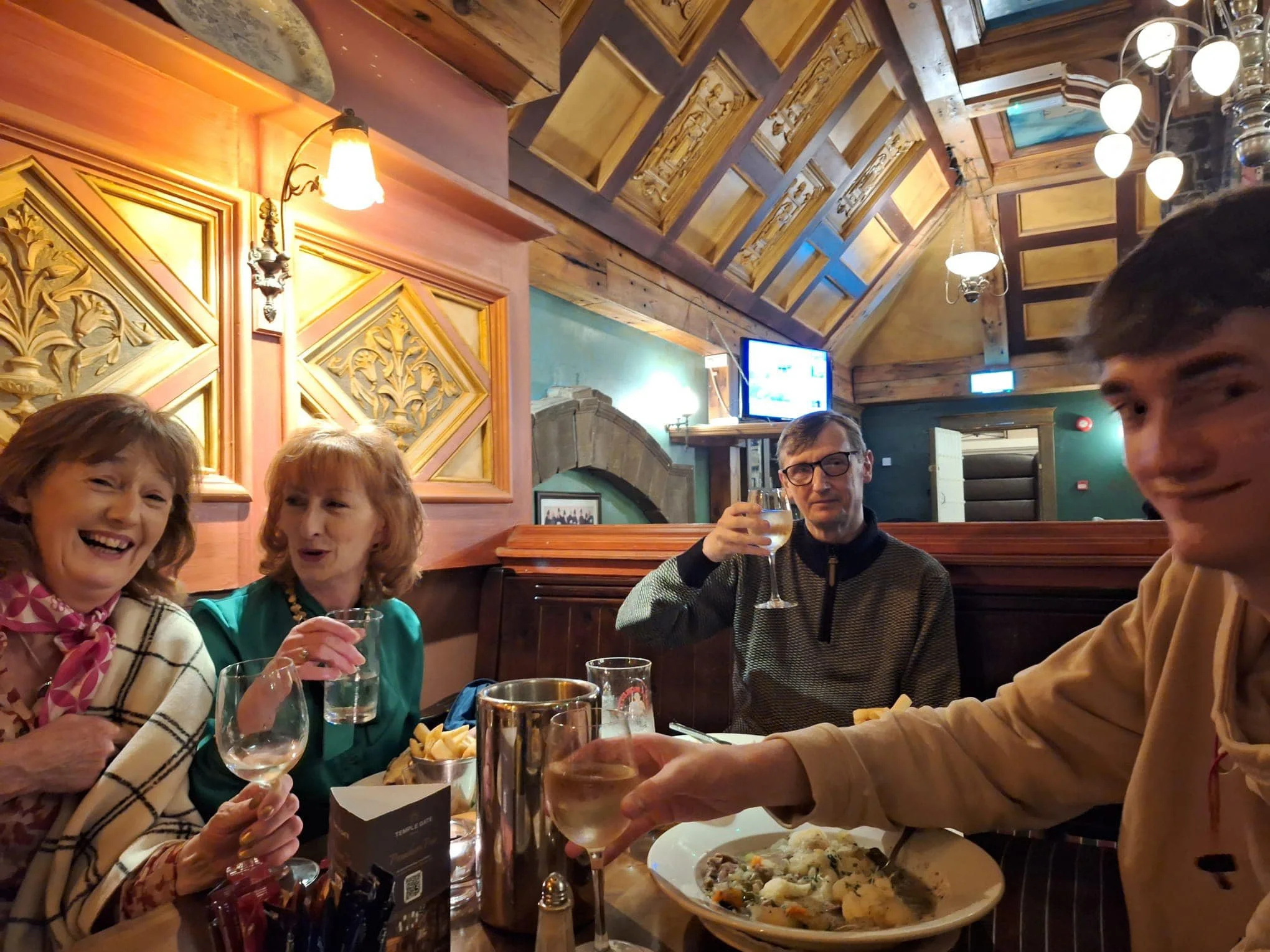 Breda and friends at a table in a cozy restaurant, smiling and holding glasses of wine or water, with a plate of food and fries on the table to celebrate her son's birthday. The restaurant has warm lighting, wooden decor, and a TV in the background.