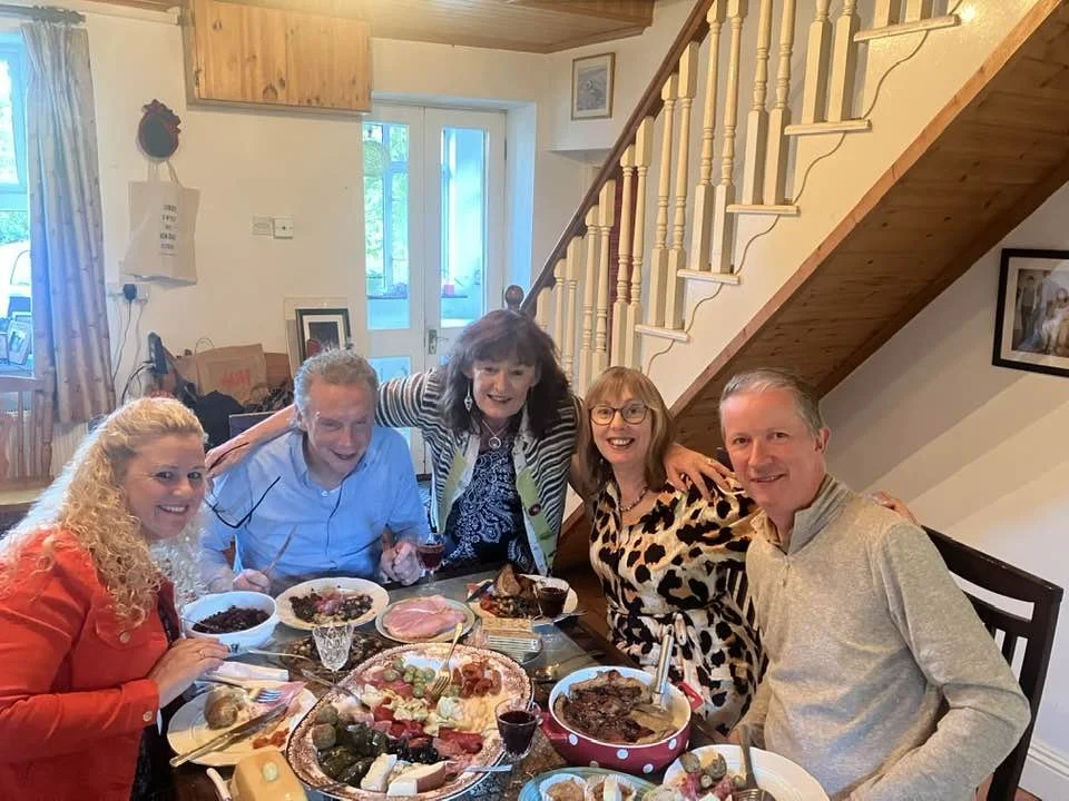 Breda and friends sitting around a dining table with various foods, smiling and enjoying a meal in a cozy home with wooden accents.