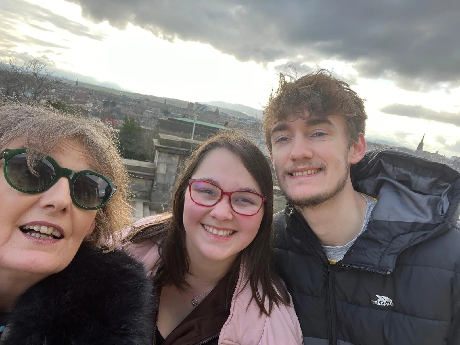 Breda, son and his girlfriend taking a selfie on a rooftop in Edinburgh with a cityscape and cloudy sky in the background.