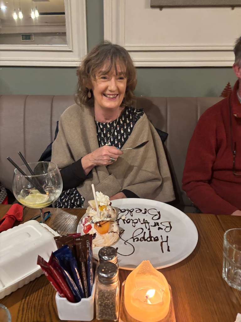 Breda celebrates her birthday with a surprise dessert at a restaurant with friends. There is a large plate with a dessert, a glass of wine, and a lit candle on the table.