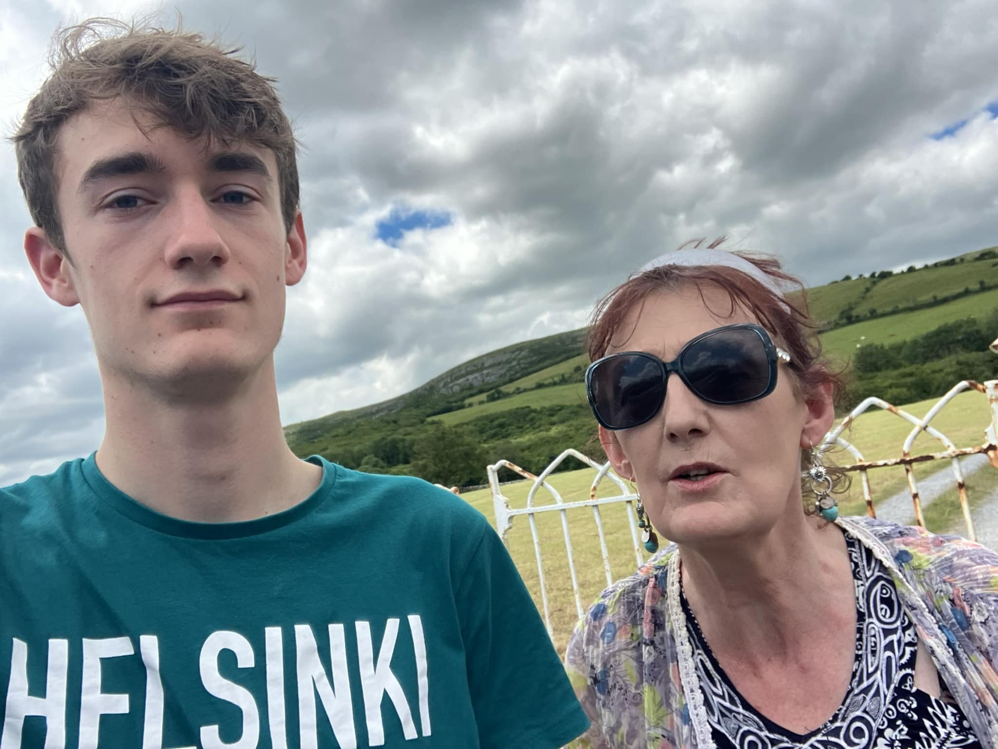 Breda and her son taking a selfie outdoors with a green hillside, cloudy sky, and rusted fence in the background.