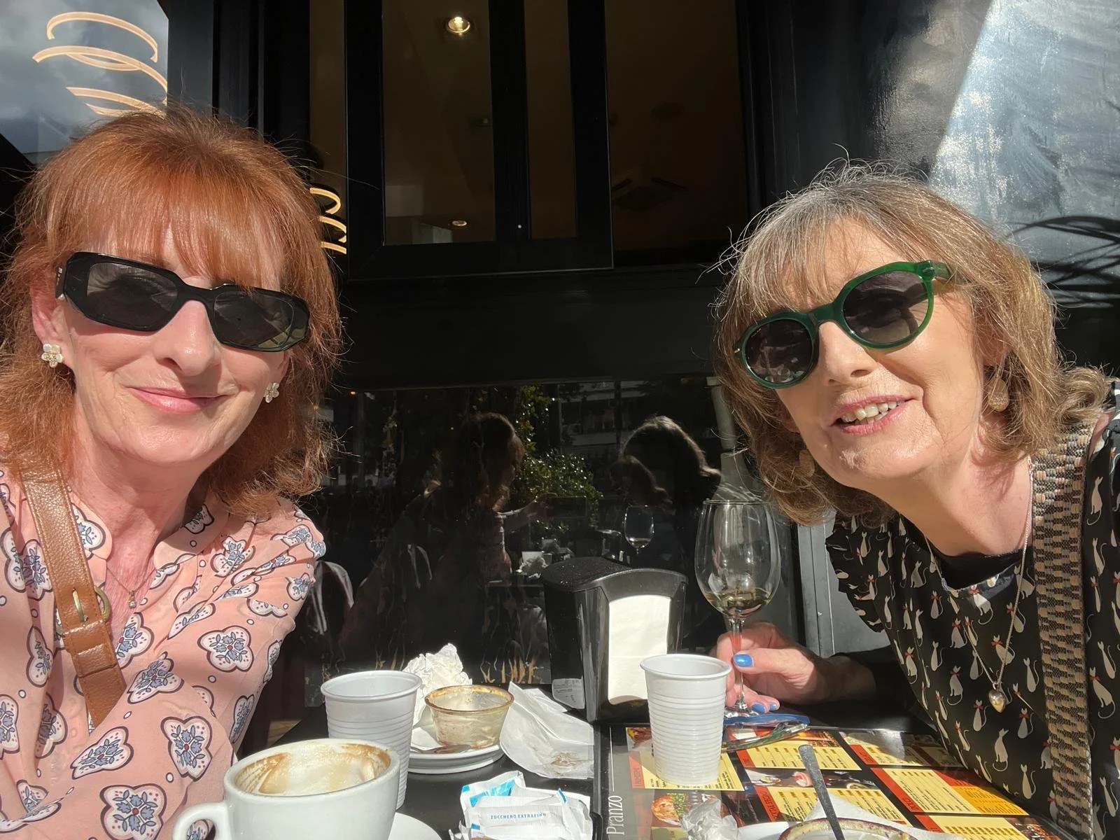Breda and her best friend with sunglasses enjoying drinks at an outdoor restaurant table with dishes and empty cups.