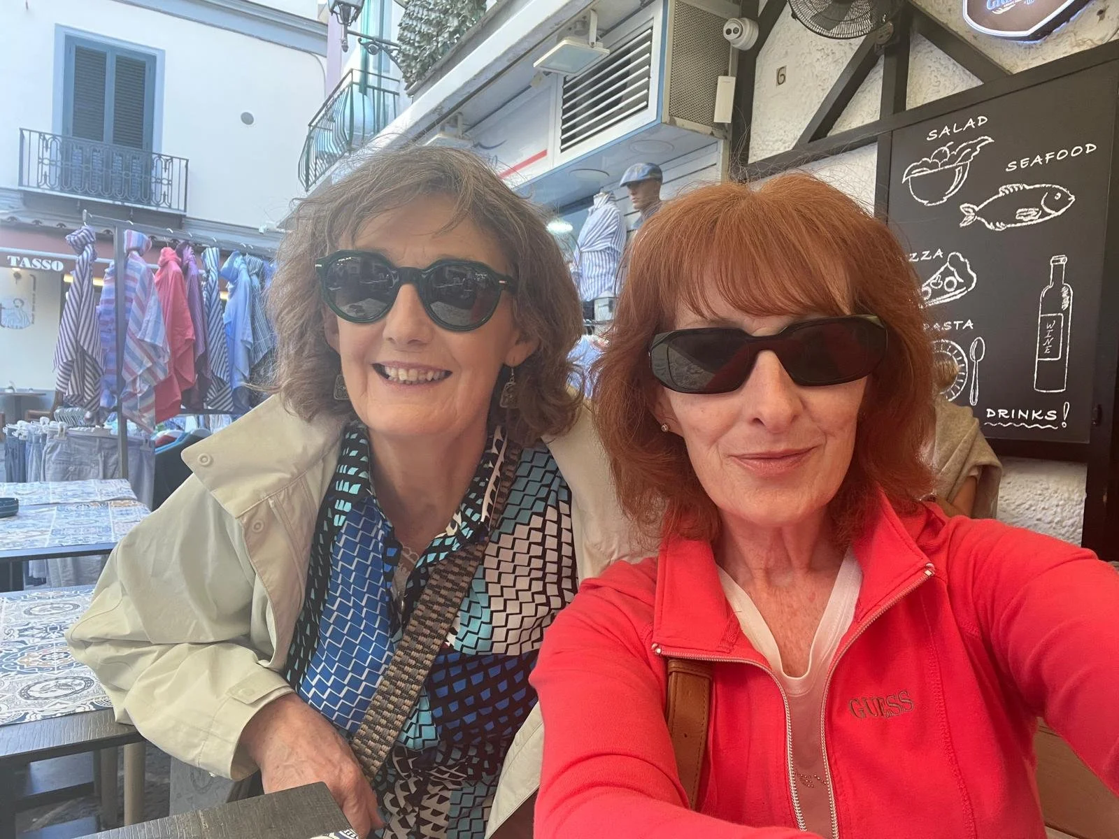 Breda and her best friend wearing sunglasses taking a selfie at a cafe in Italy with outdoor seating, colorful striped umbrellas in the background, and a blackboard menu with illustrations of food and drinks.