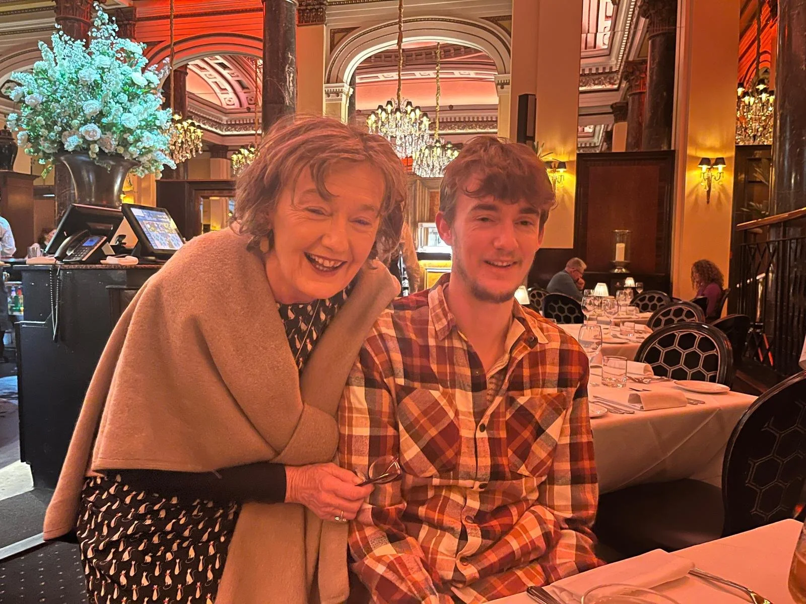 Breda with a beige shawl smiling and leaning towards her son in a plaid shirt sitting at a table in an elegant restaurant