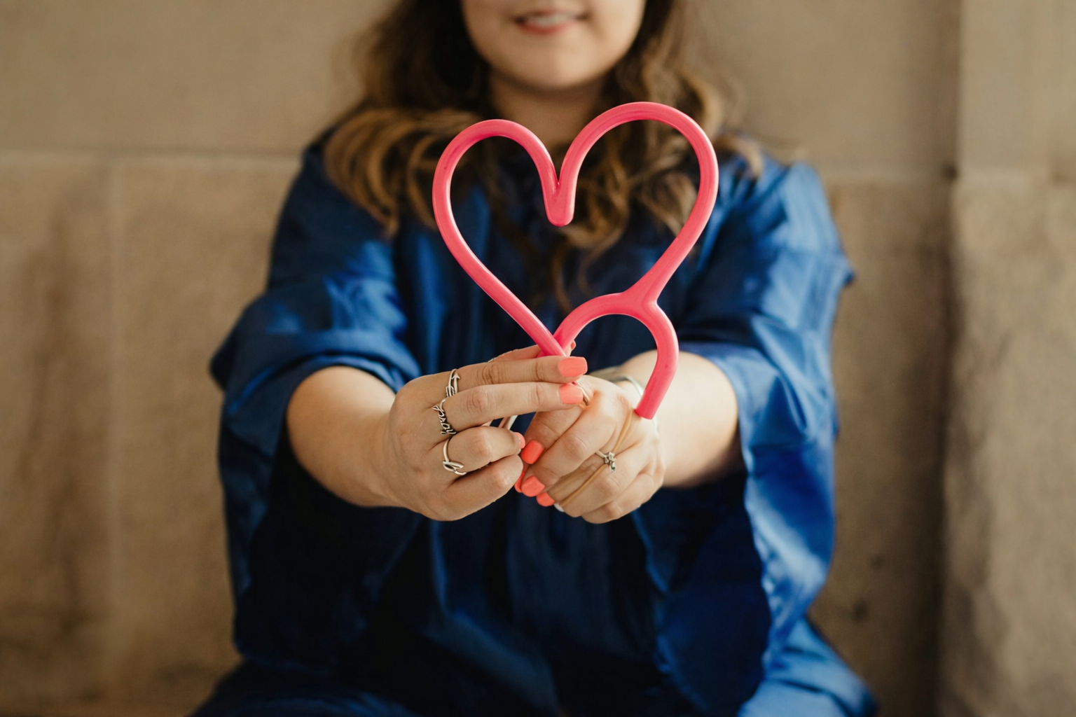 A woman with long brown hair, wearing a blue satin top, holds a pink heart-shaped balloon sculpture outwards.