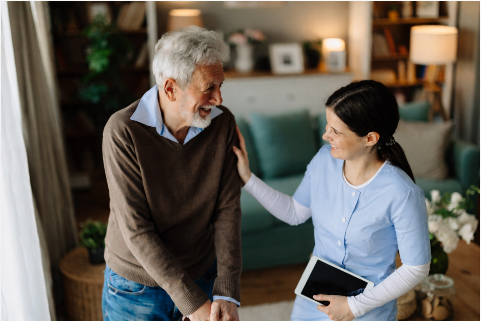 A nurse smiling and interacting with an elderly man in a cozy living room, holding a tablet device.