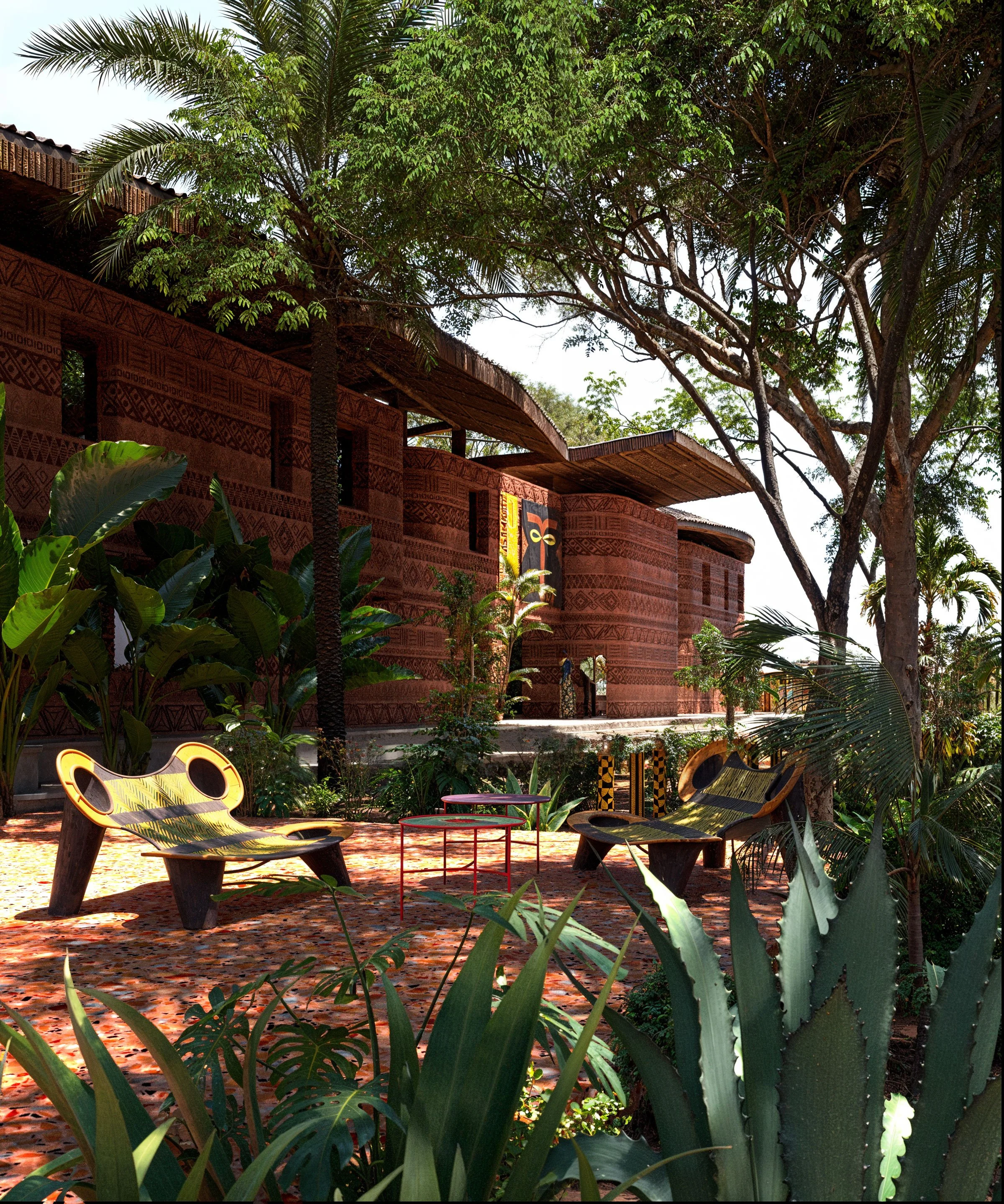 Outside view of a traditional building with intricate carvings surrounded by lush tropical plants and trees, including large agave and palm plants, with two modern benches and two small tables in the foreground.