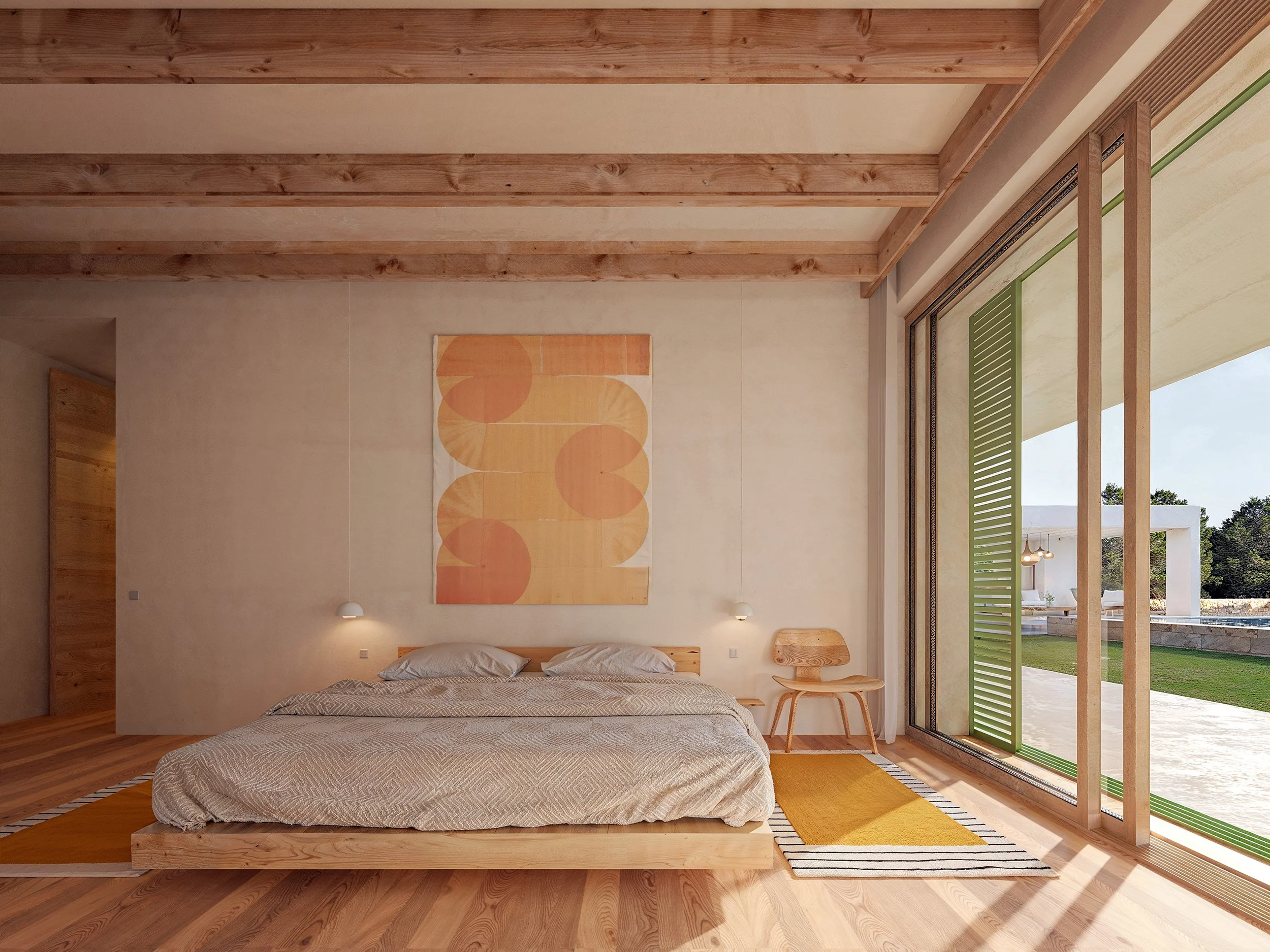 A minimalist bedroom with a low wooden bed, beige bedding, and two pillows. Above the bed hangs an abstract orange and yellow artwork. To the right, large sliding glass doors open to a green outdoor space, allowing natural light to fill the room. The