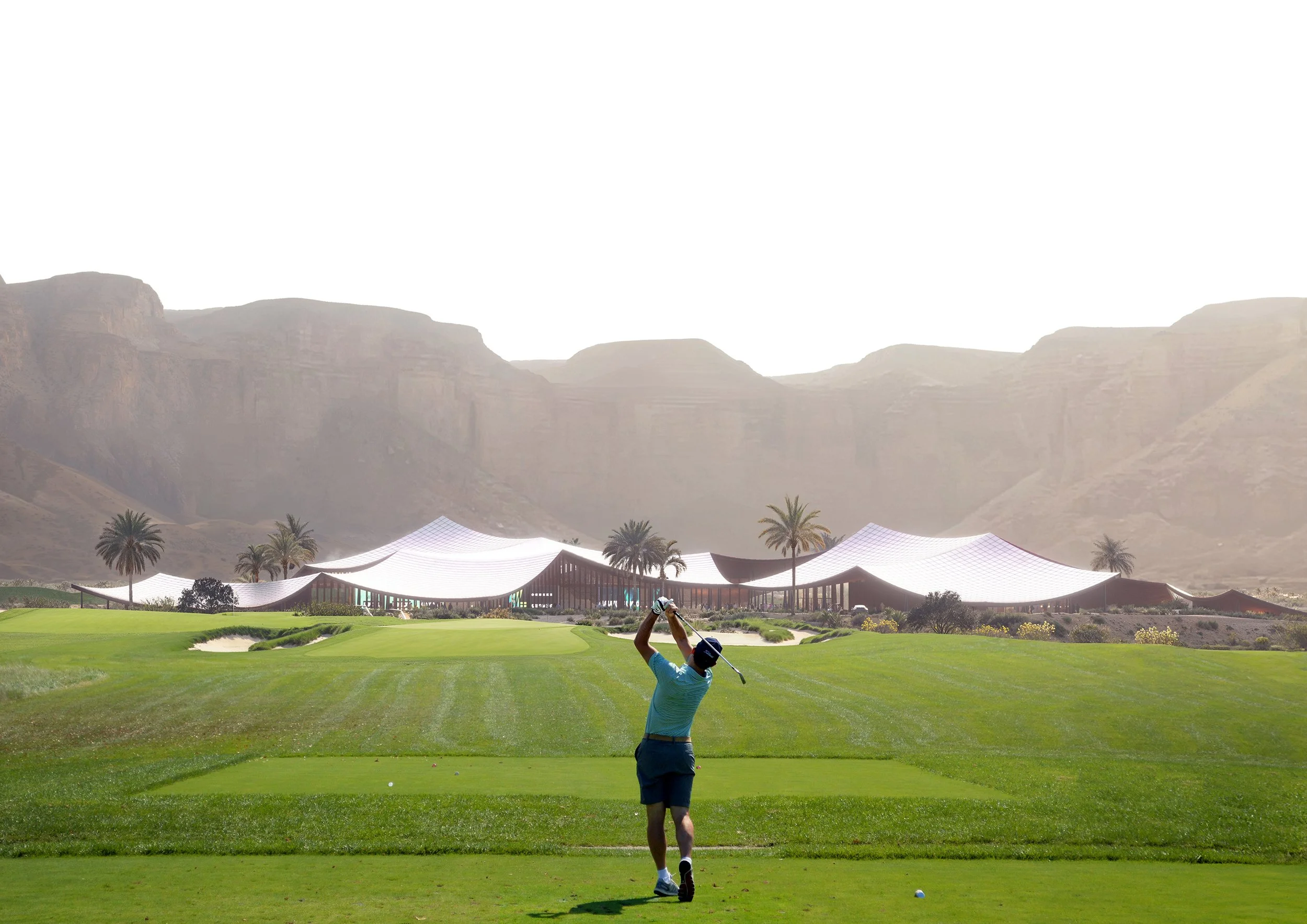 A person playing golf on a green golf course with a modern building and palm trees in the background, with mountains behind.