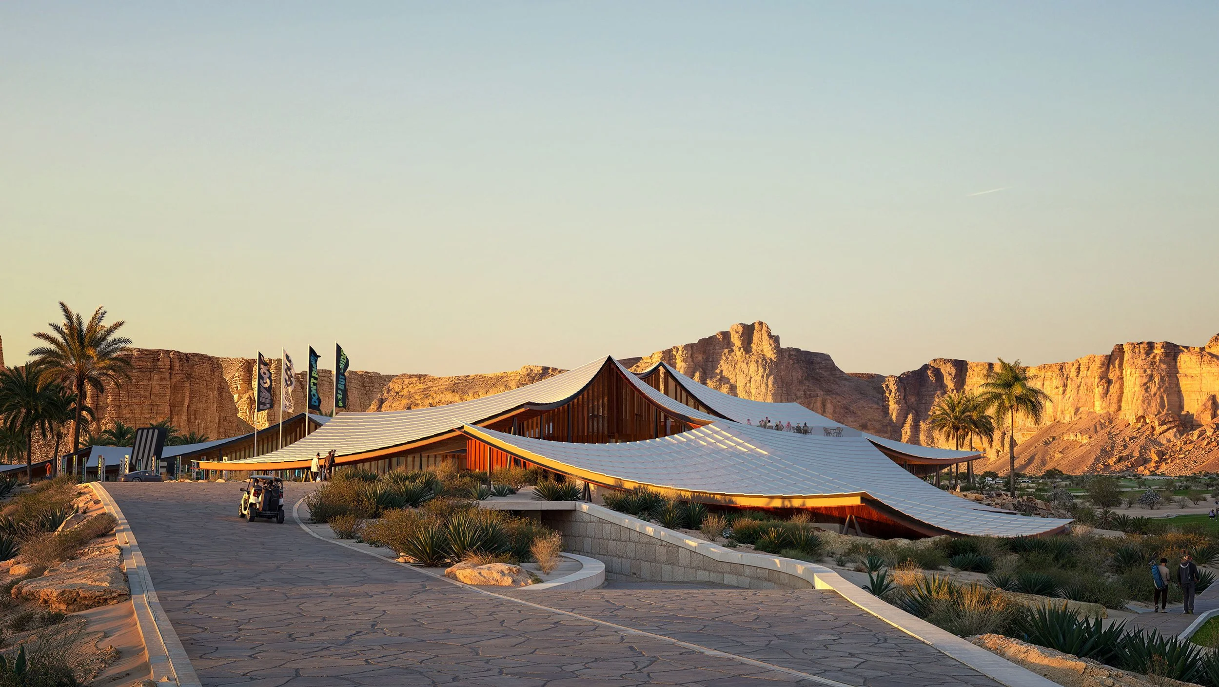 Modern building with curved roof in desert landscape, surrounded by palm trees and rocky cliffs at sunset.