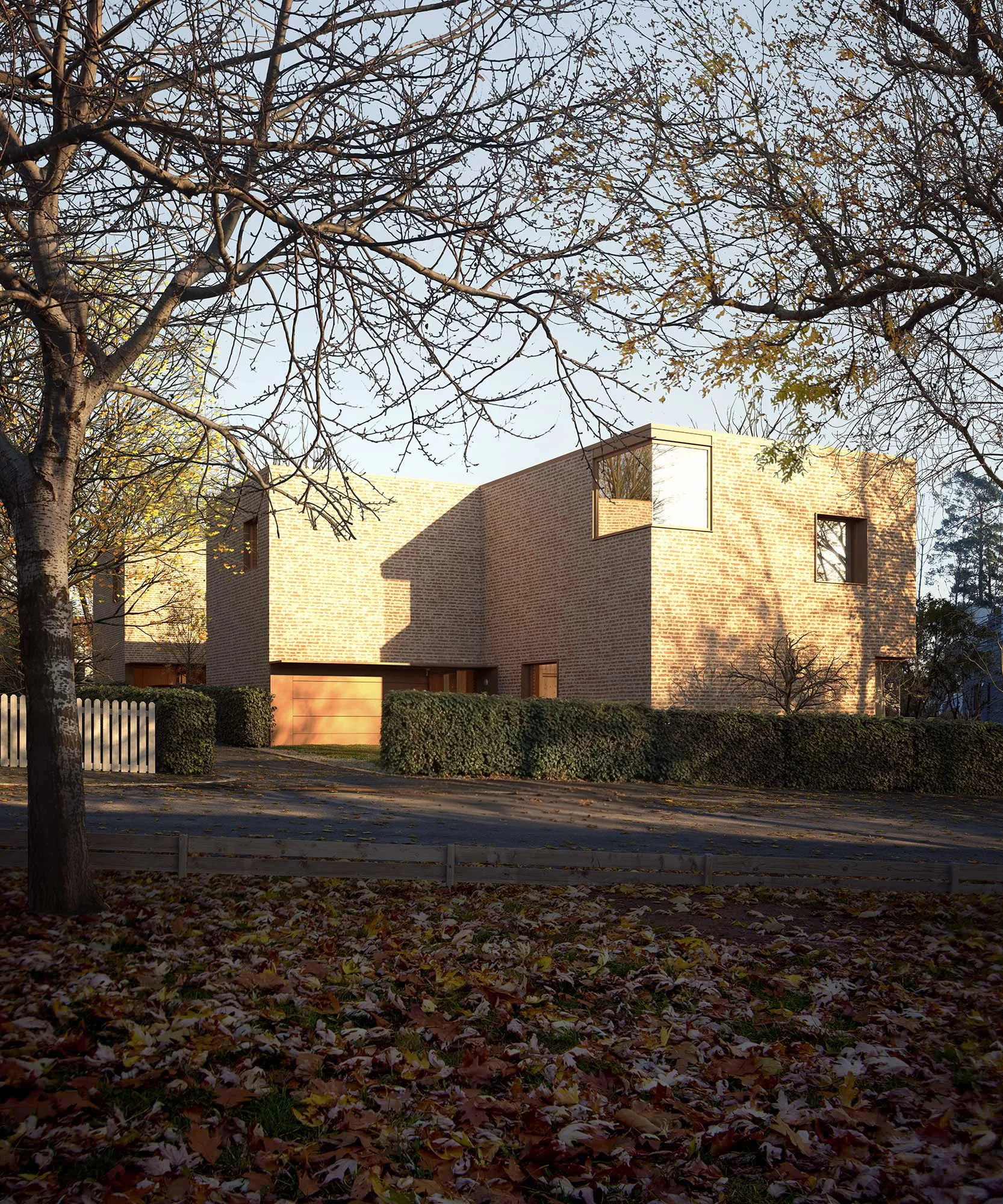 A modern brick house with large windows, surrounded by fallen autumn leaves and trees with sparse branches.