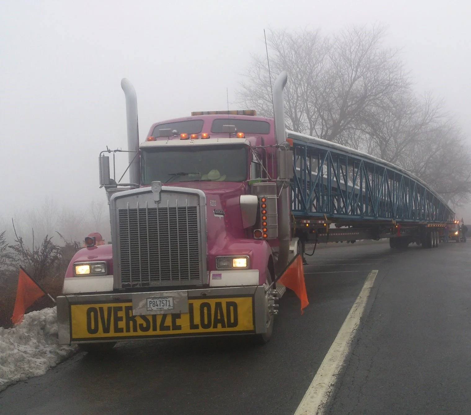 A large semi-truck with a pink cab and a yellow "Over Size Load" sign on the front, carrying a blue metal structure on a flatbed trailer, parked on the side of a foggy road with leafless trees in the background.