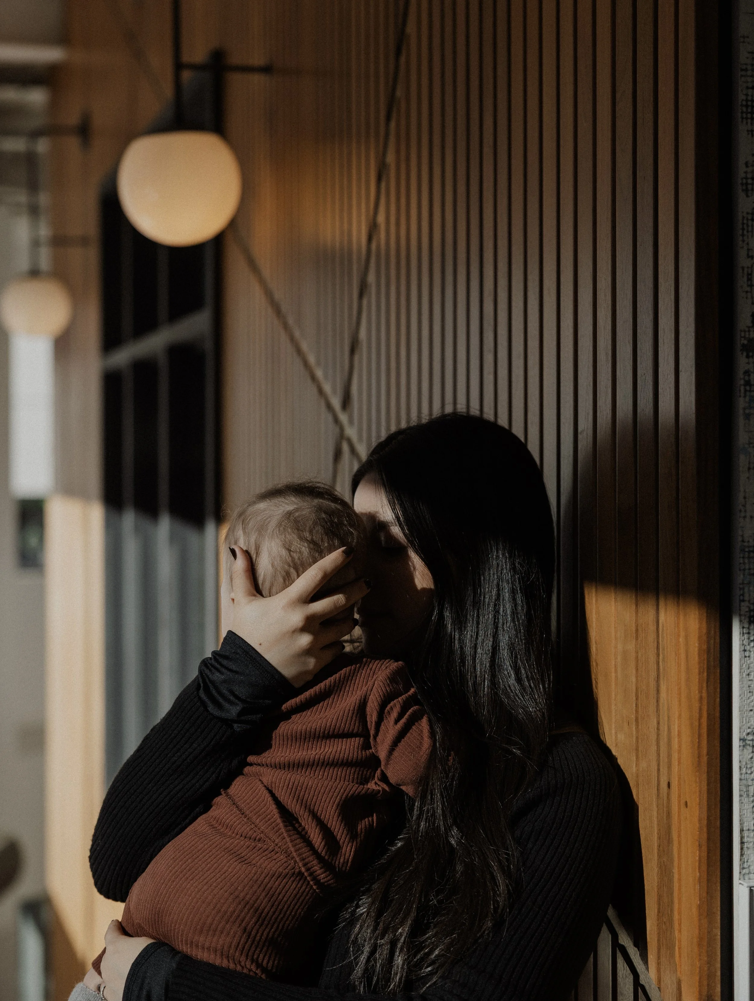 A woman with long black hair hugging and kissing a child with blonde hair in an indoor setting with wooden wall paneling and warm lighting.