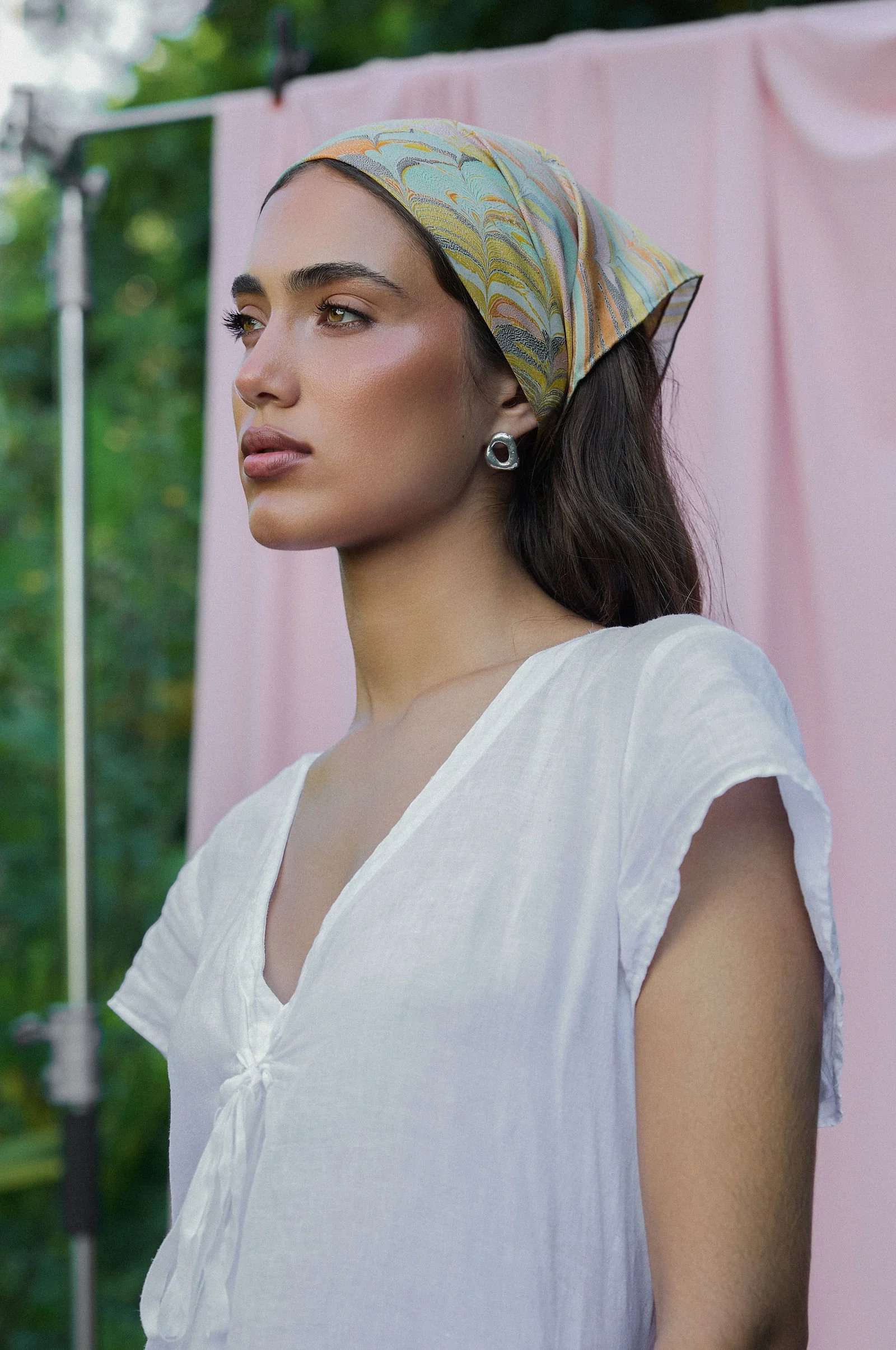 A woman wearing a patterned headscarf, silver earrings, and a white dress, looking to the side with greenery in the background and a pink cloth hanging.