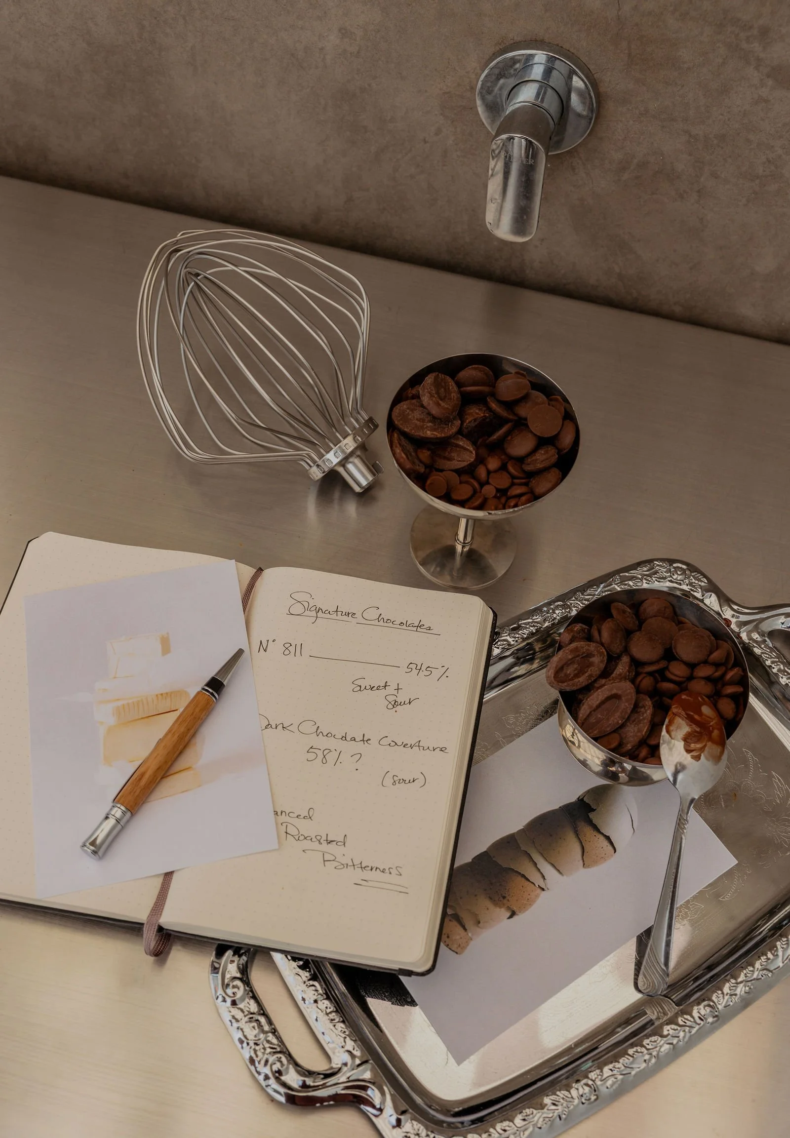 A silver tray with a sheet of paper, a pen, a notebook, and two glasses filled with chocolate chocolates and cookies, on a beige table.
