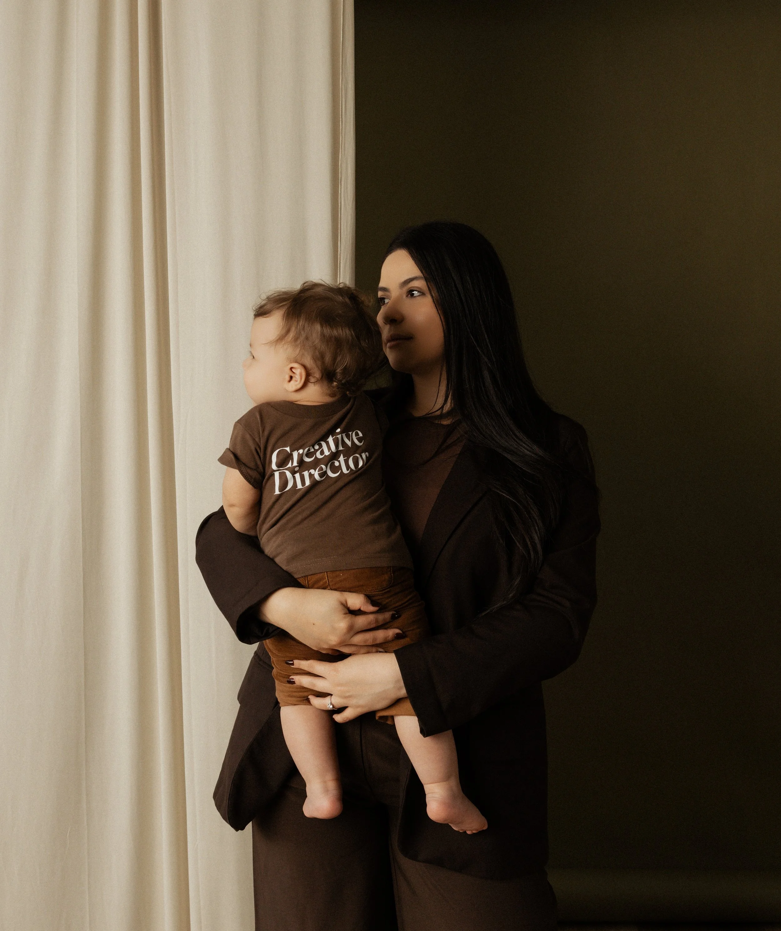 A woman with long dark hair holding a young child in her arms indoor near a dark wall and a light-colored curtain.