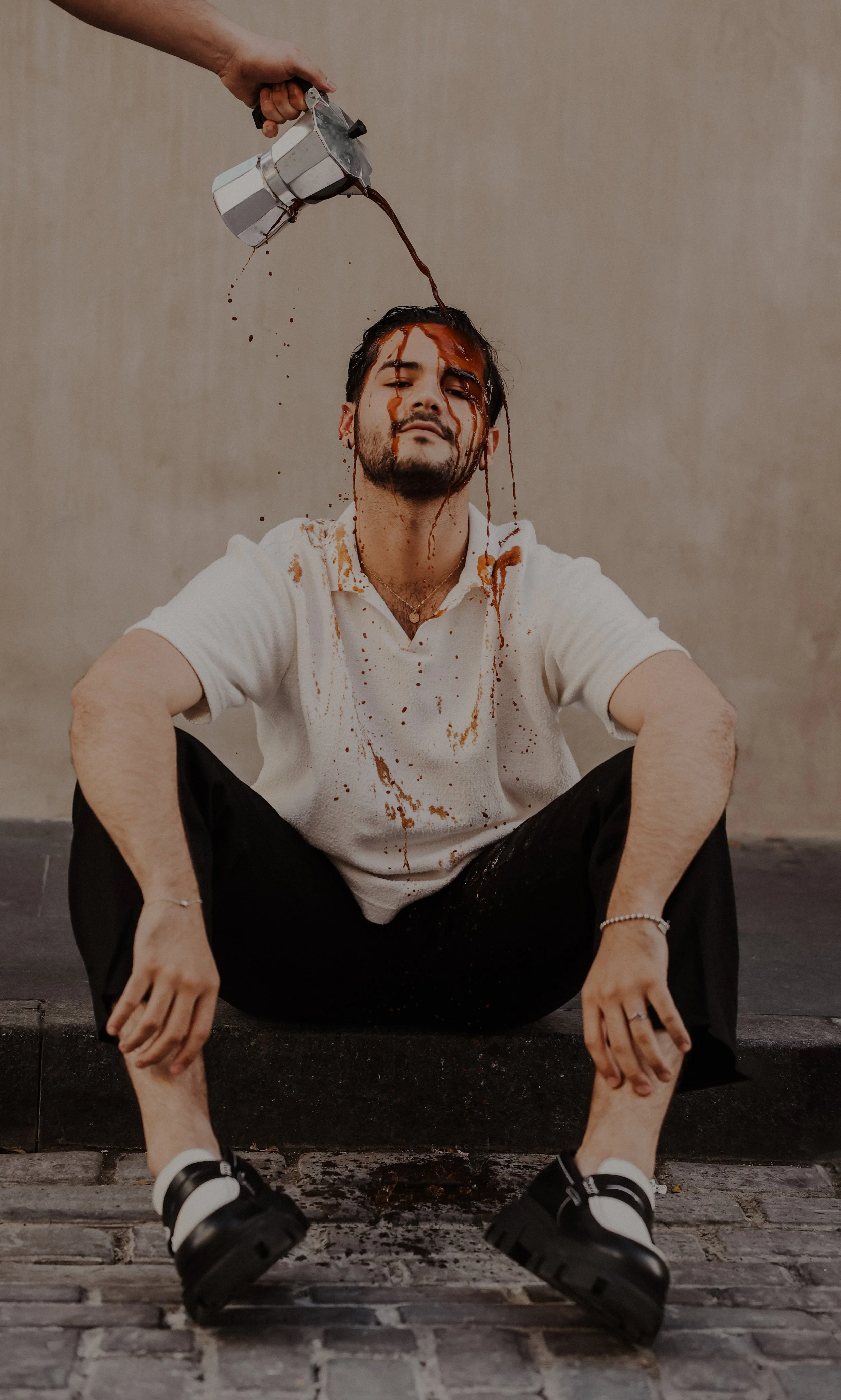 A man sitting on the ground with his knees bent and arms resting on his knees. He has dark hair and a beard, wearing a white shirt, black pants, and black sneakers. Someone is pouring coffee over his head, causing the coffee to spill down his face and shirt. The background is a plain, beige wall.